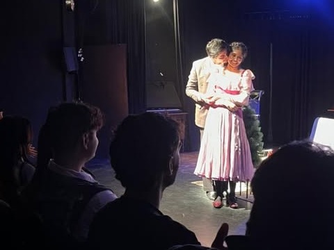 Two actors embrace under a spotlight while the audience watches